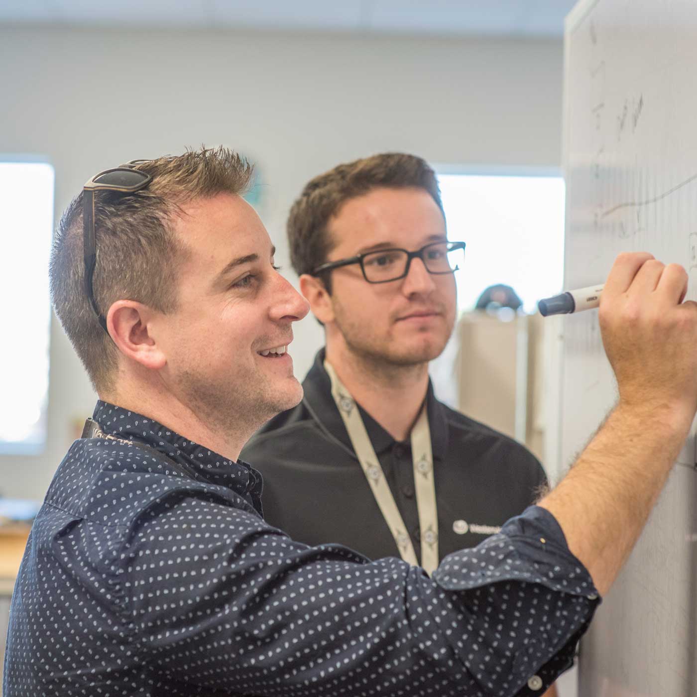 Two people standing at a whiteboard, one of them writing on it.